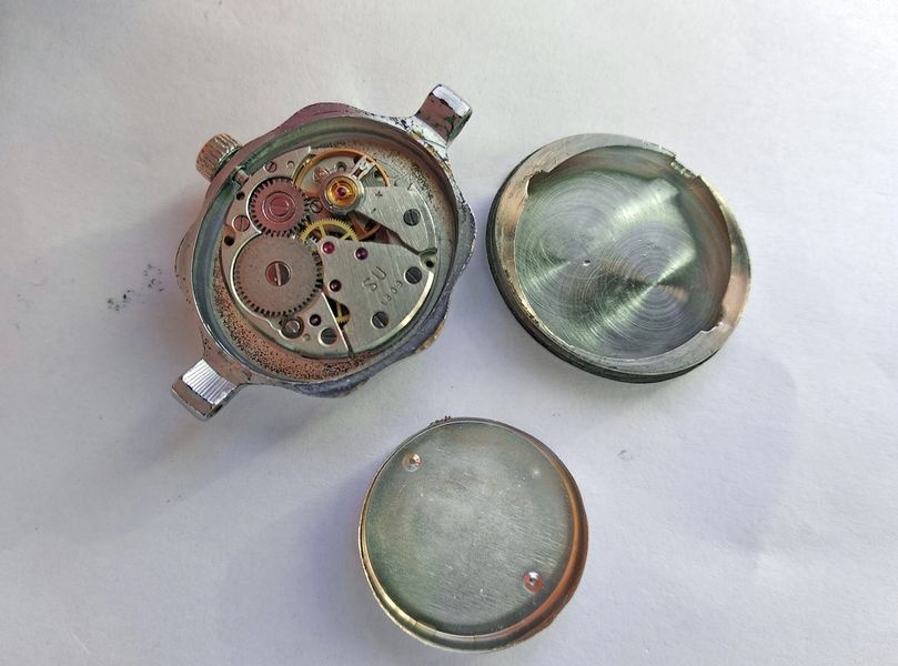 Opened Luch case with the movement in place, beside the removed case back and inner dust cover