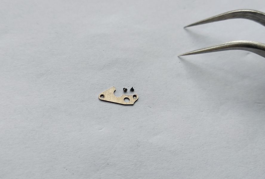 Small minute wheel bridge and its two screws lying on the bench beside tweezers