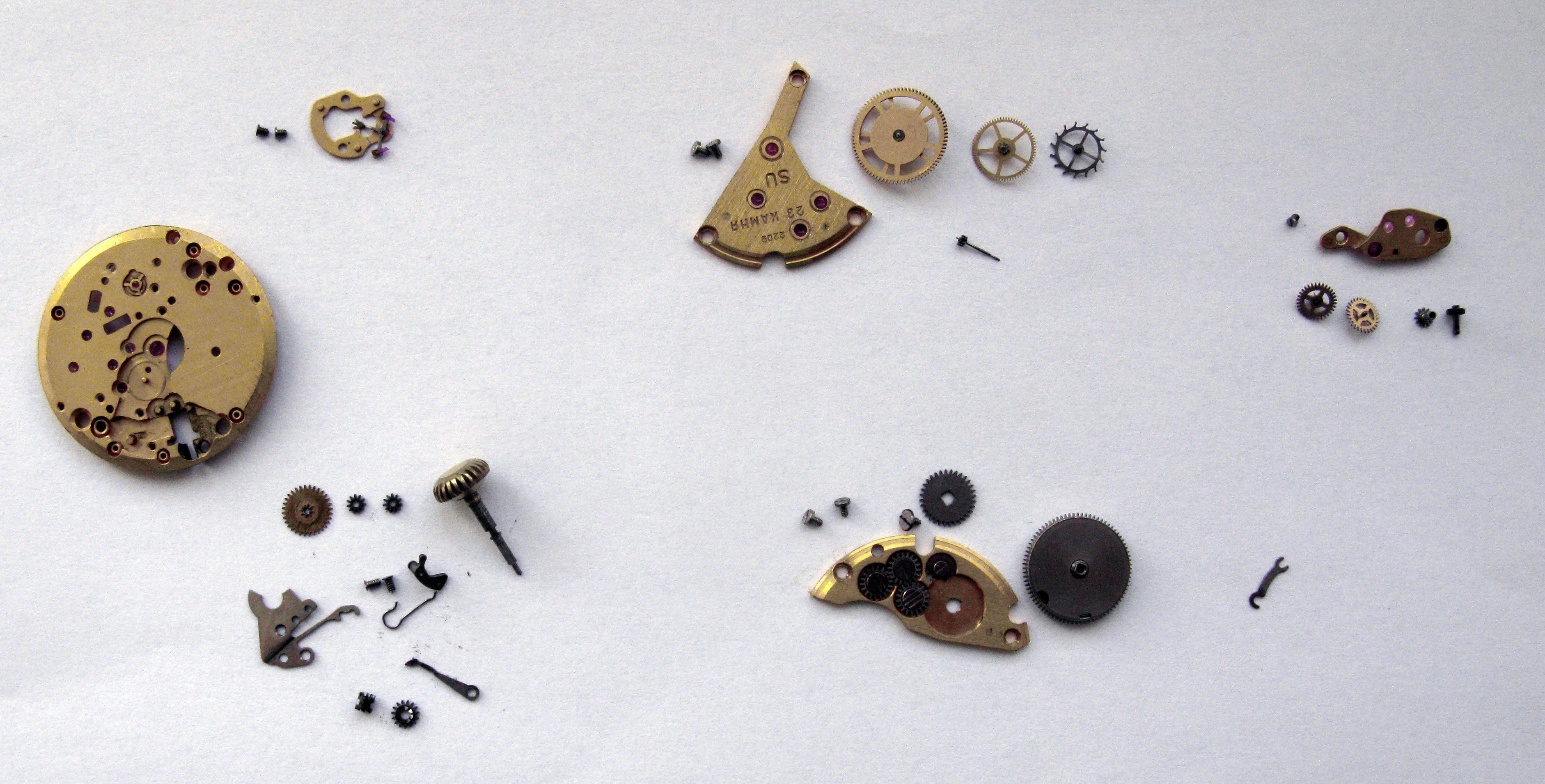 All movement parts laid out in rows on a white sheet, including plates, bridges, wheels, barrel and steelwork.