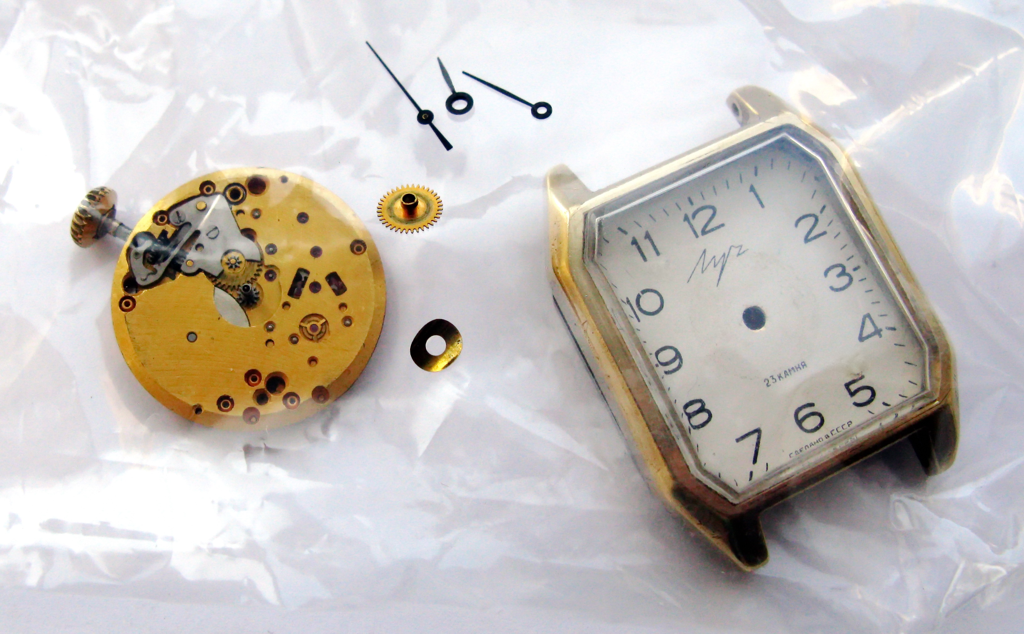 The dismantled Luch wristwatch as received, with the gilt rectangular case and dial beside the loose mainplate, hands and winding components.
