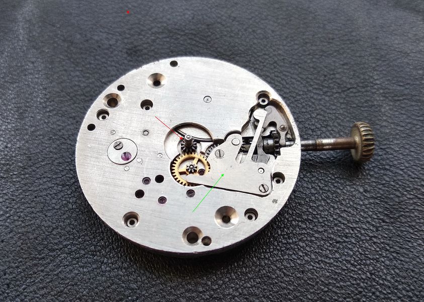 Cannon pinion at the movement center marked with a red arrow, with the keyless-works spring cover indicated by a green arrow