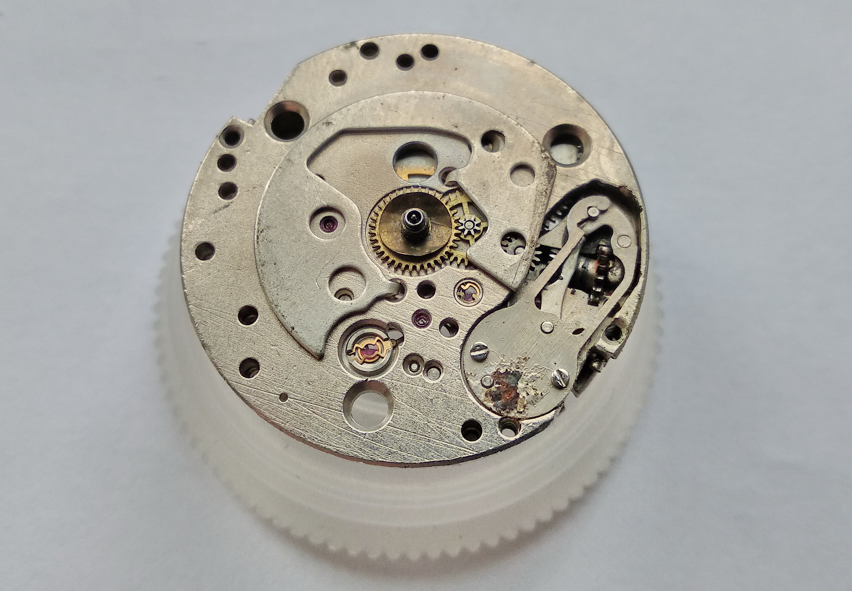 Under-dial view showing the hour wheel with spring washer, minute wheel, setting wheel, and a rusty keyless cover spring
