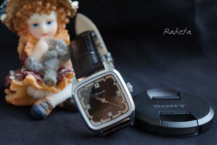 The chrome version staged with a porcelain doll and a black lens cap on a dark surface