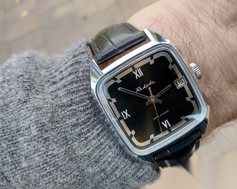 The chrome version with a black Roman-numeral dial worn on the wrist over a grey sweater