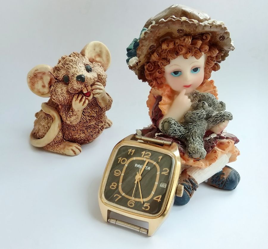 The finished gold Raketa with Arabic-numeral dial staged on a white background beside porcelain doll and mouse figurines
