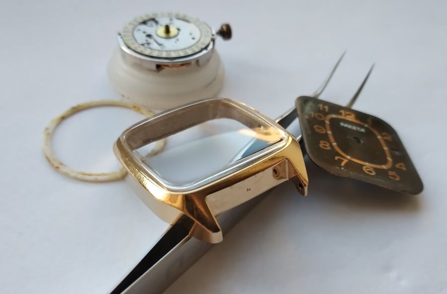 The gold-plated case with crystal, the white plastic locating ring, the running movement, and a gilt dial held in tweezers