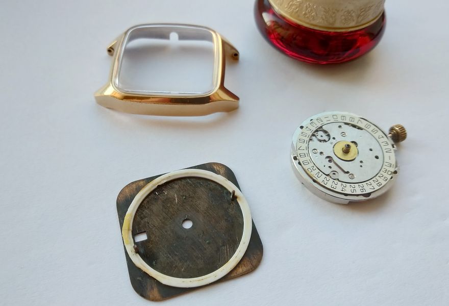 The gilt dial face-down with the white plastic locating ring fitted to its back, beside the gold case and movement