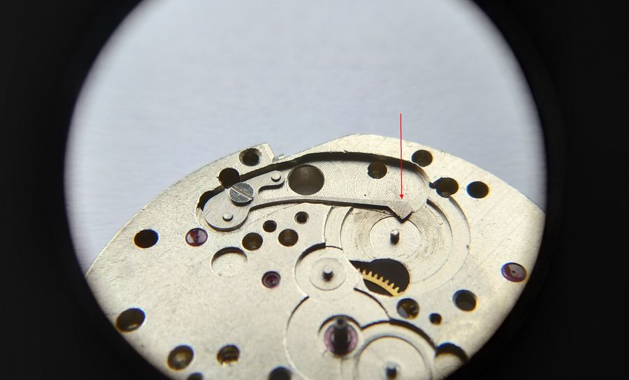 A loupe view of the plate with the slender accumulating spring in place, a red arrow marking its hooked beak