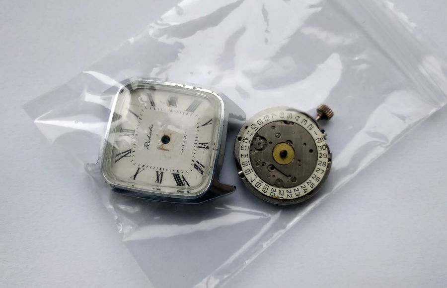 A small clear plastic bag holding a square Raketa watch case and a loose movement, set on a white surface