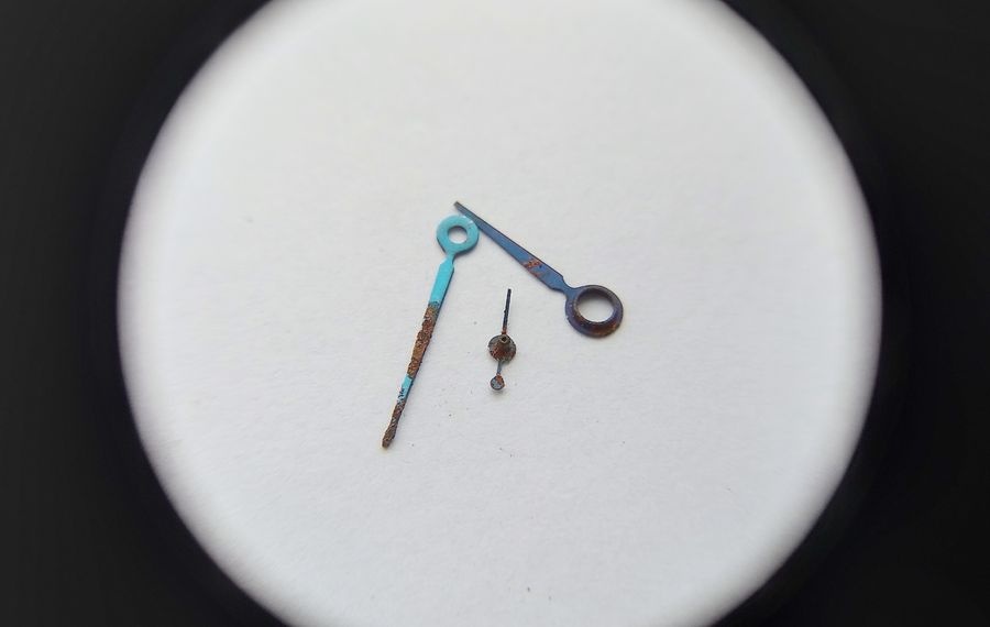 Three removed Vostok watch hands through loupe showing blue steel hour minute and seconds hands with surface oxidation
