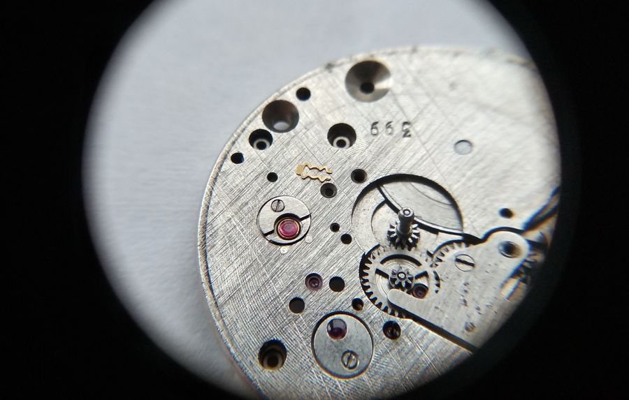 Vostok 2605 dial side through loupe showing lyre spring being fully removed from main plate before bouchon can be extracted